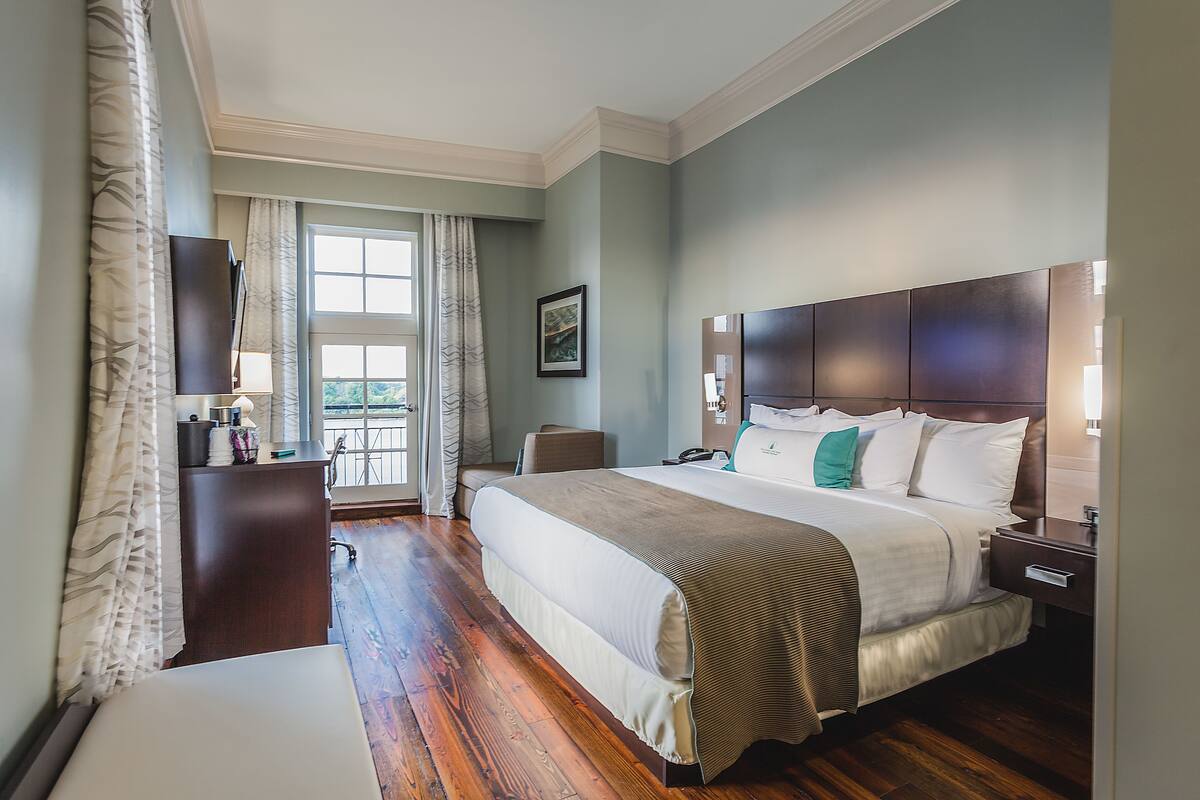 A bedroom with a large bed, wooden flooring, a dresser, a nightstand, and a window with curtains.