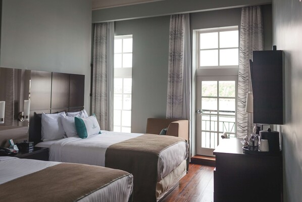 A hotel room with two beds, a desk, a TV, and large windows with curtains.
