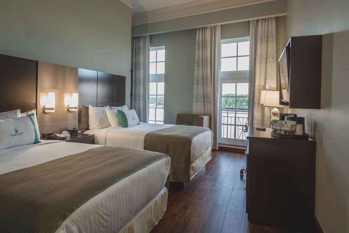 A hotel room with two beds, a desk, a chair, a TV, and a balcony with curtains.