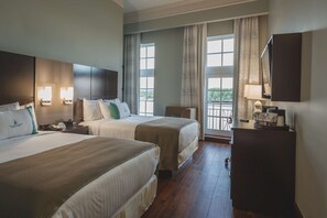 Ett hotellrum med två sängar, ett skrivbord, en stol, en TV och en balkong med gardiner.