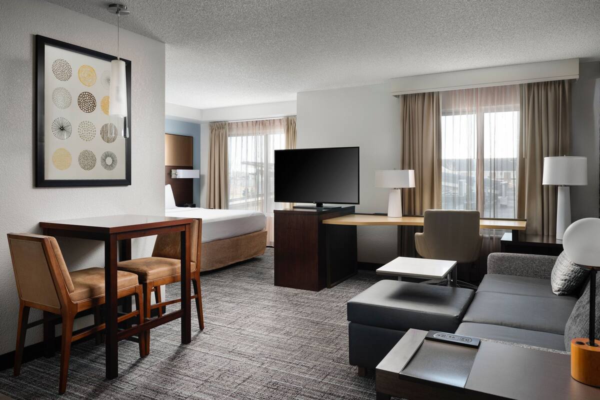 A hotel room with a bed, a desk with a TV, a sofa, and a dining area with chairs.