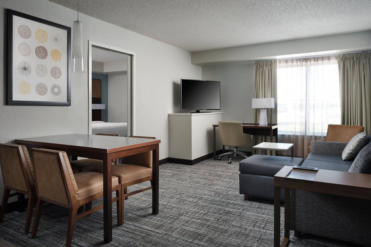A hotel room with a dining area, a sofa, a TV, and a window with curtains.