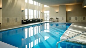 Una piscina interior con agua cristalina y azul, rodeada de paredes con azulejos y grandes ventanales que dejan entrar luz natural.