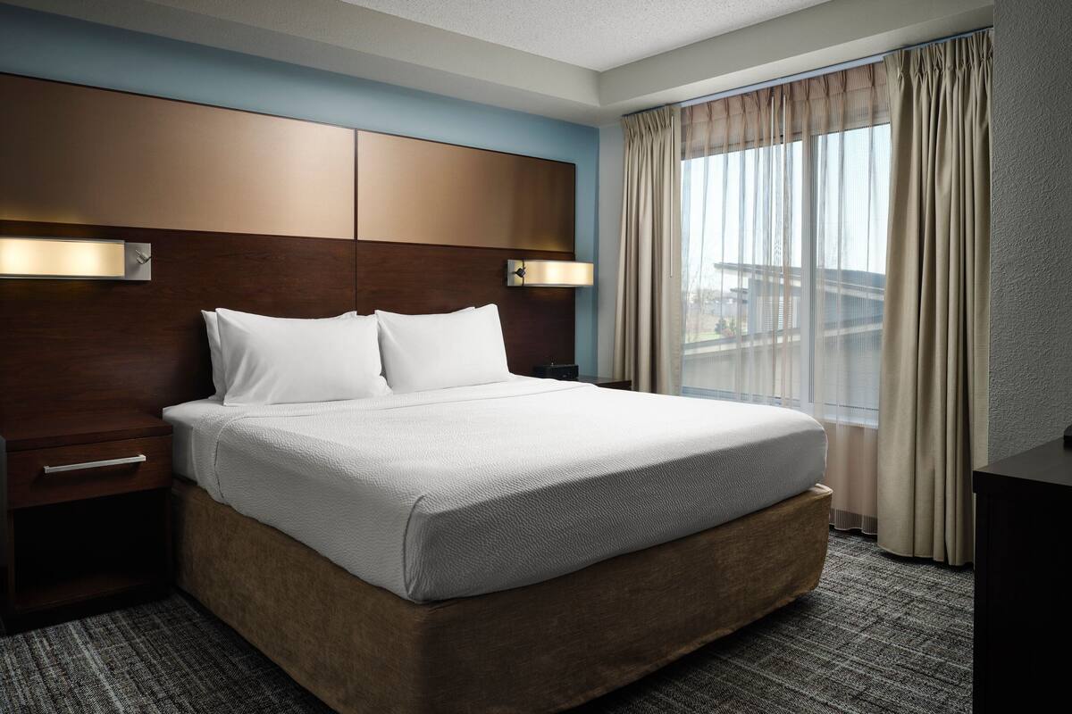 A hotel room with a large bed, a nightstand, a wall-mounted light, and a window with curtains.