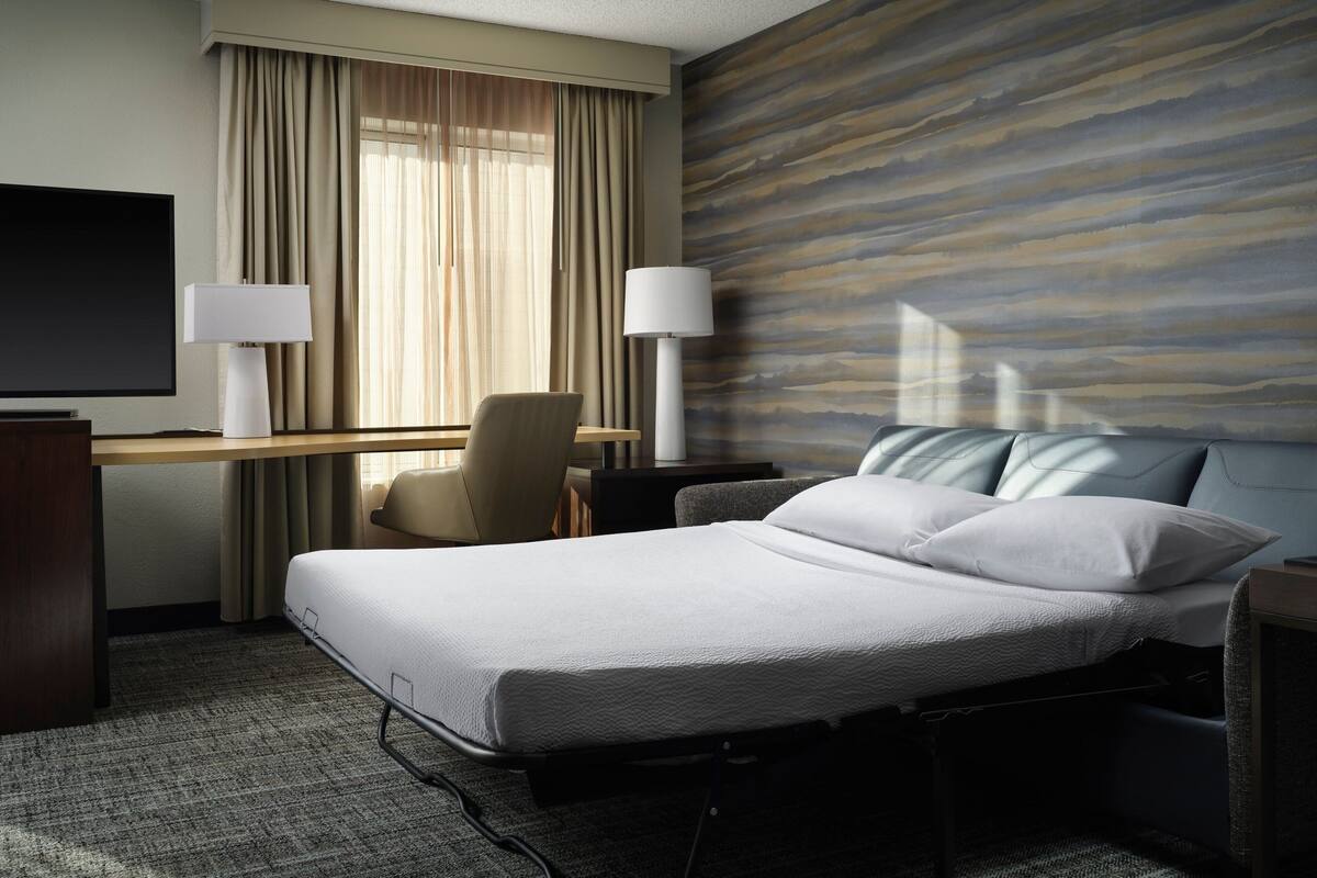 A hotel room with a bed, a desk, a chair, two lamps, and a TV.