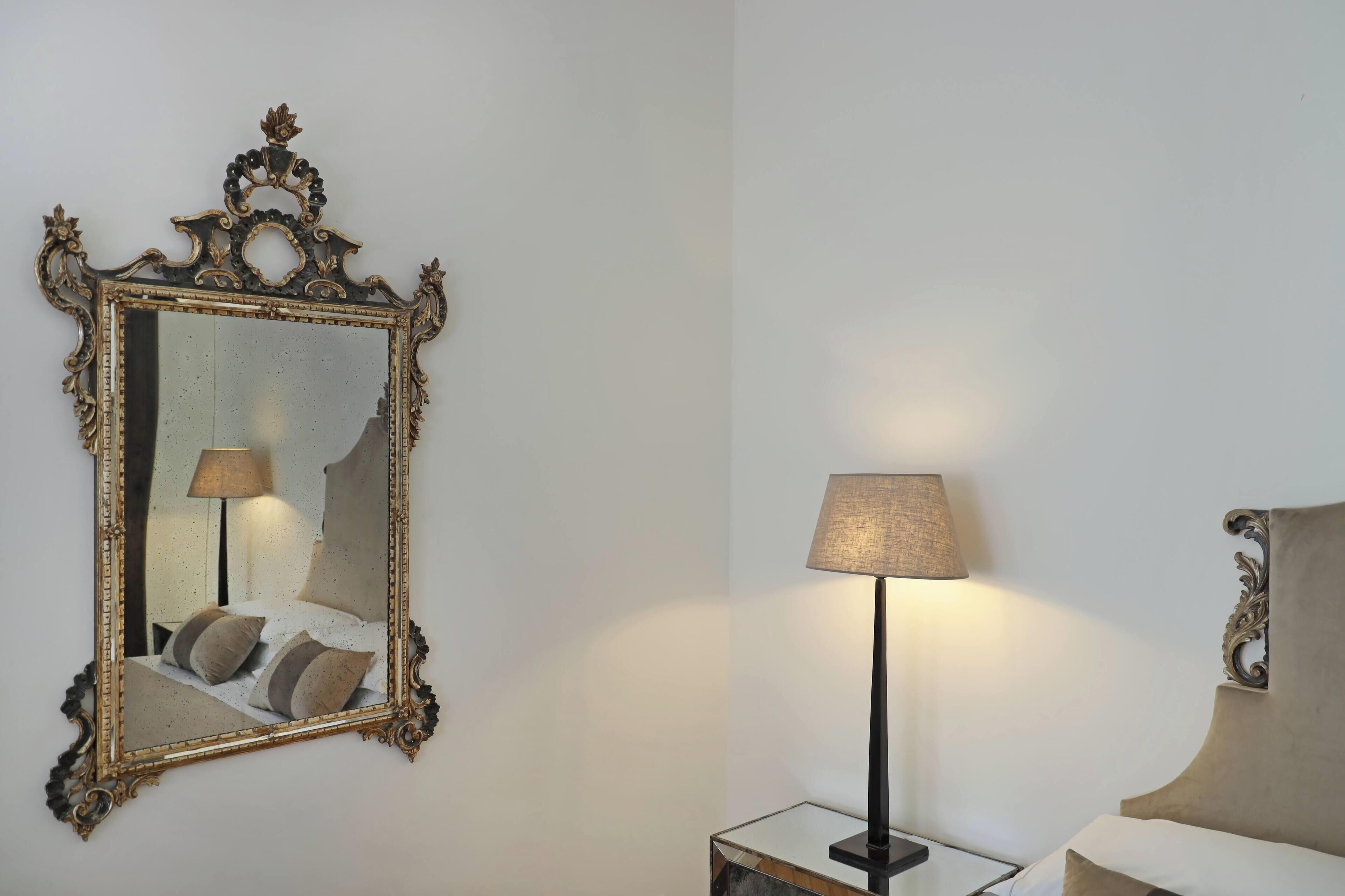 A bedroom with a large ornate mirror, a bedside lamp, and a bed with pillows.