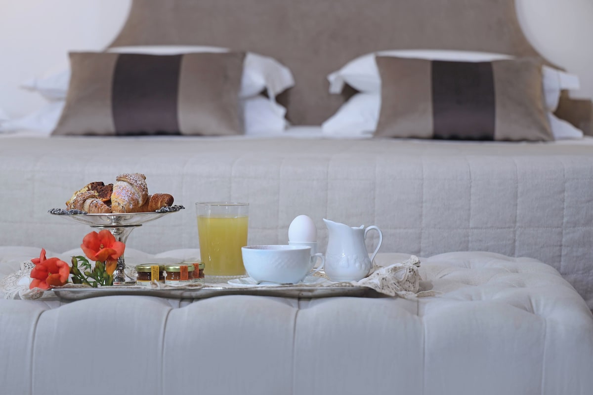 A breakfast setup on a cushioned bench with pastries, a glass of orange juice, a bowl, a pitcher, and jars of jam.