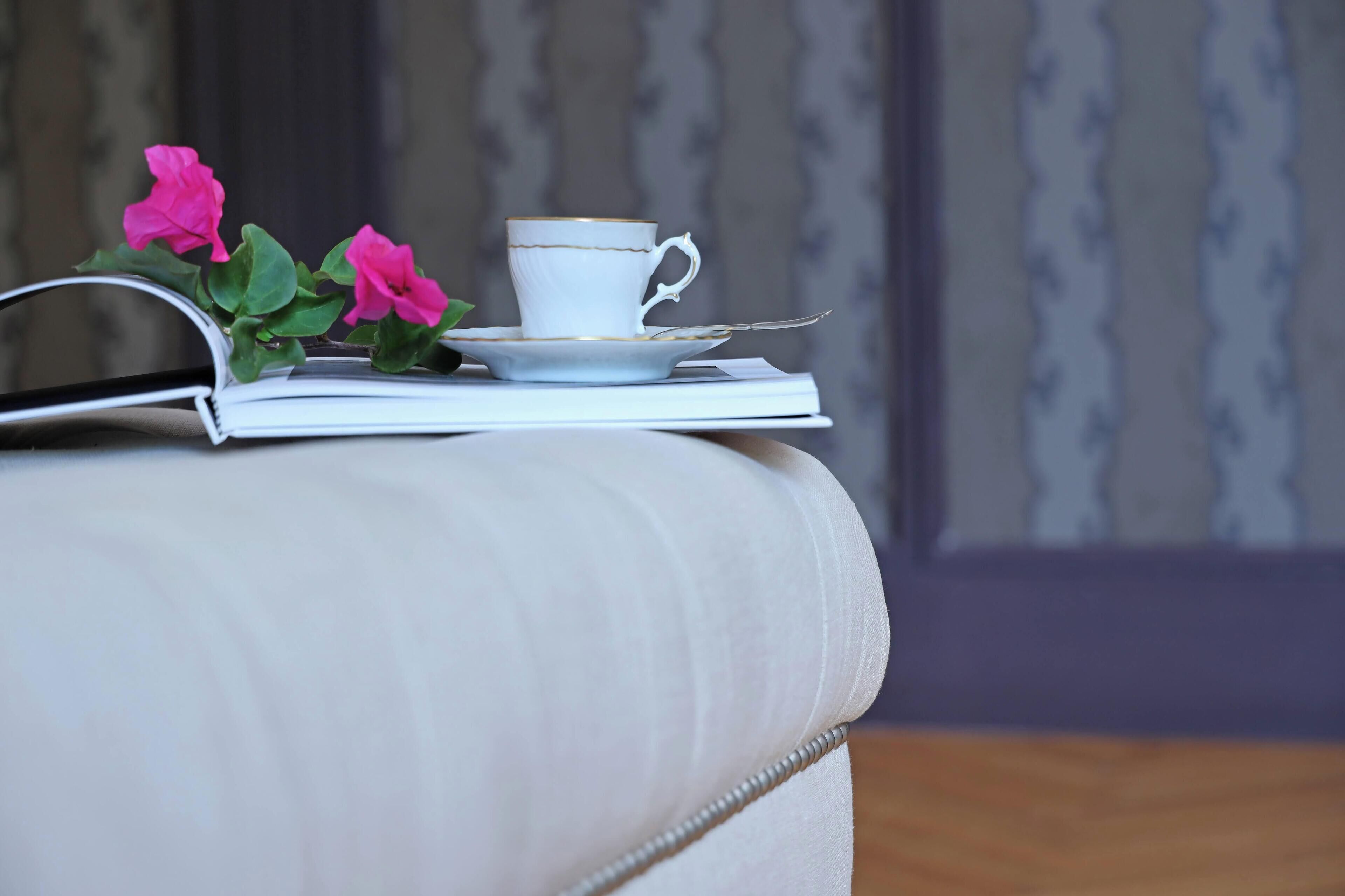 A white cushioned bench with a cup and saucer on top, accompanied by pink flowers and a book.