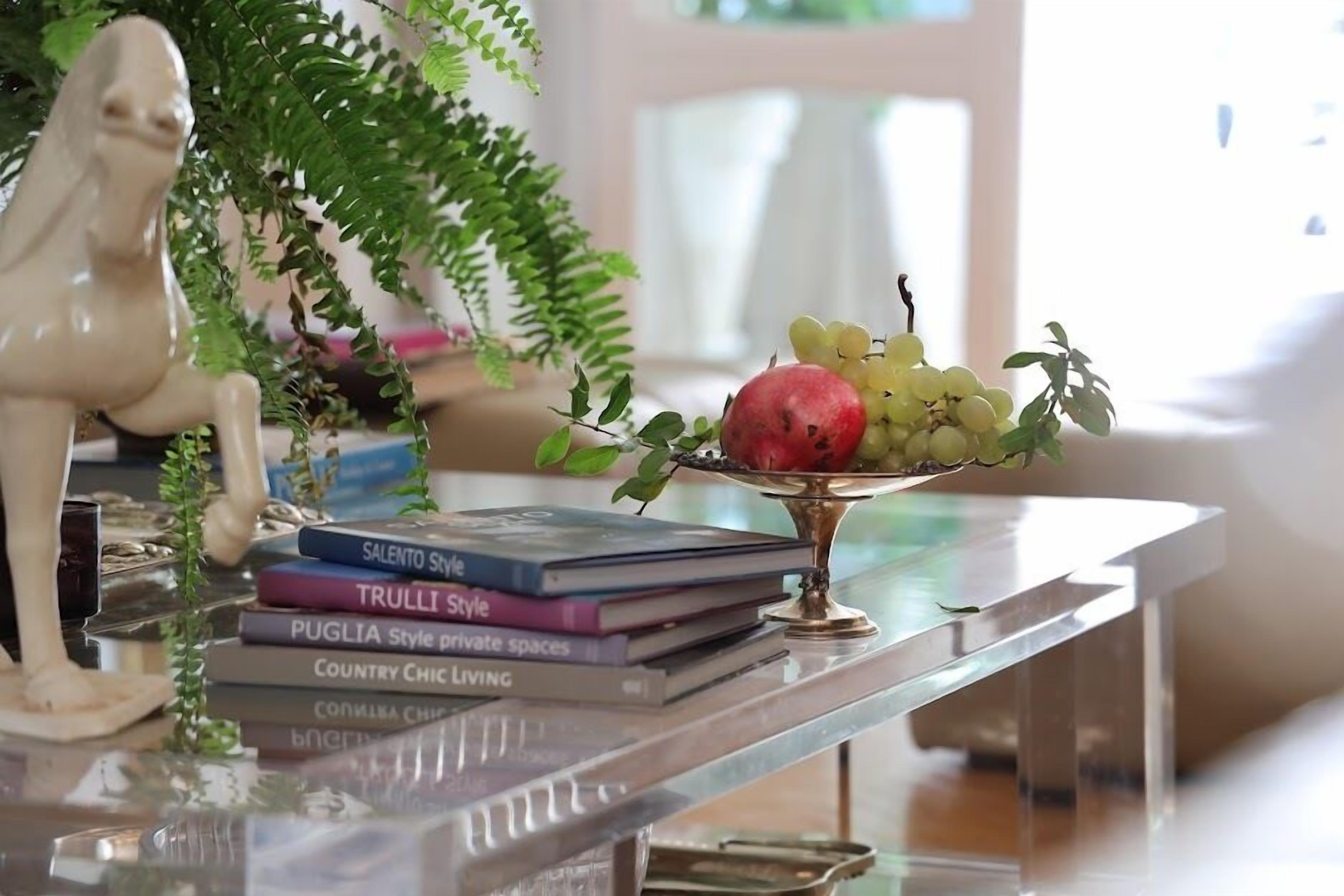 Una mesa de vidrio transparente con un cuenco metálico decorativo que contiene frutas y una pila de libros.