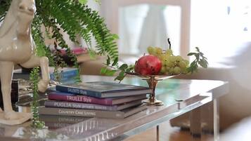 Una mesa de vidrio transparente con un cuenco metálico decorativo que contiene frutas y una pila de libros.