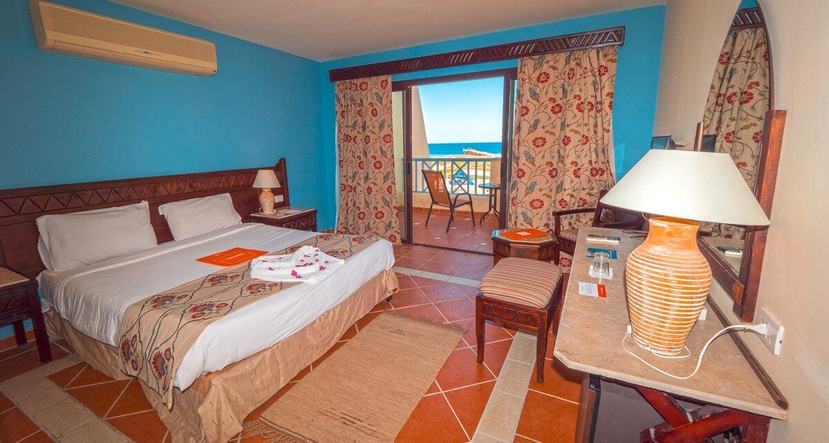 Habitación de hotel con cama, escritorio con lámpara, silla, balcón con vistas y ventana con cortinas.