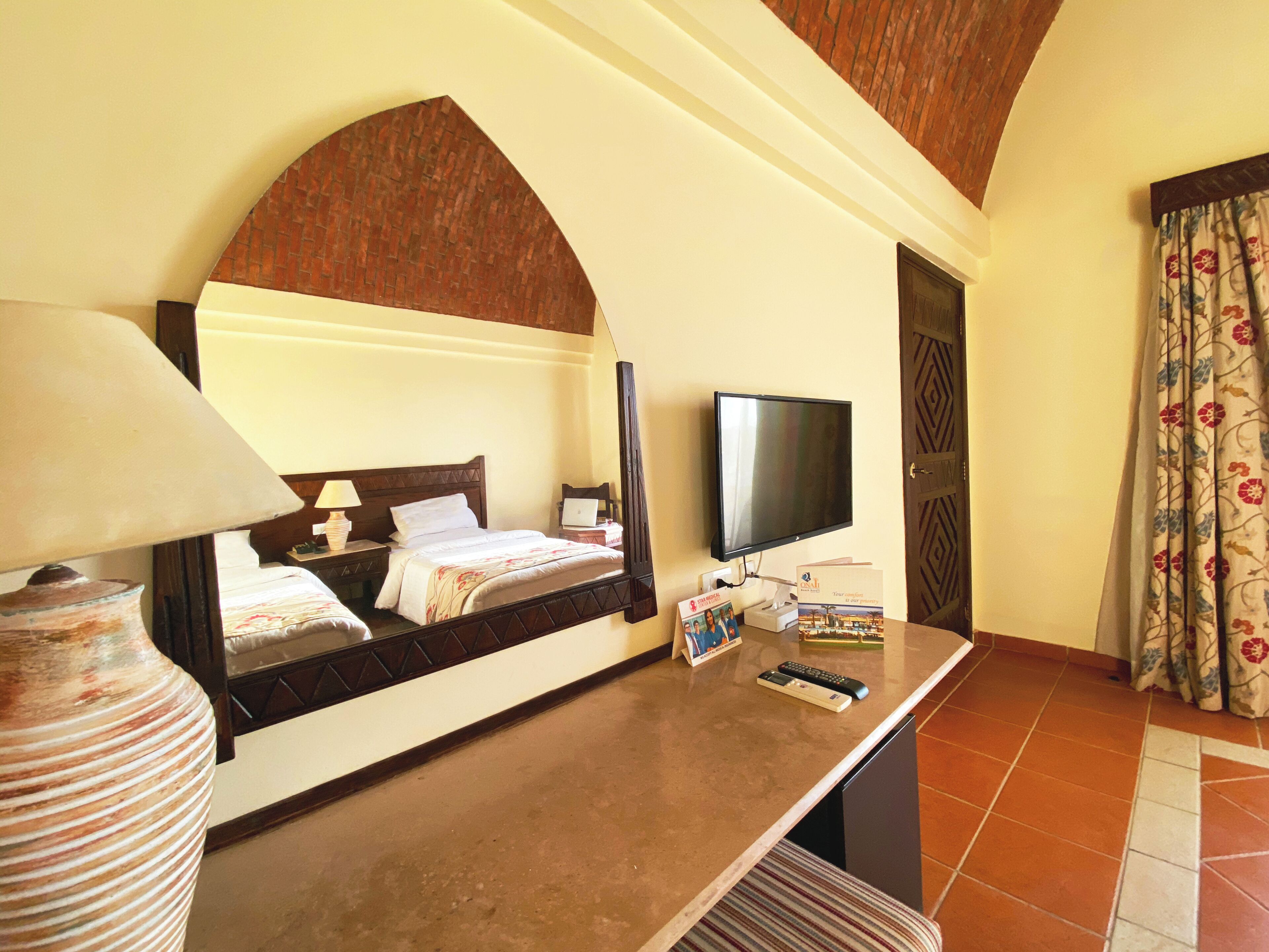 Habitación de hotel con cama, televisor, escritorio con lámpara, un florero y espejo.
