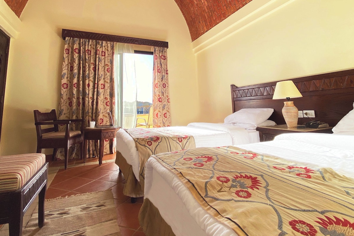 Ein Hotelzimmer mit Ziegeldecke, zwei Betten mit Blumenmuster-Bettwäsche, einem Holz-Nachttisch, einem Sessel und Blick ins Freie.