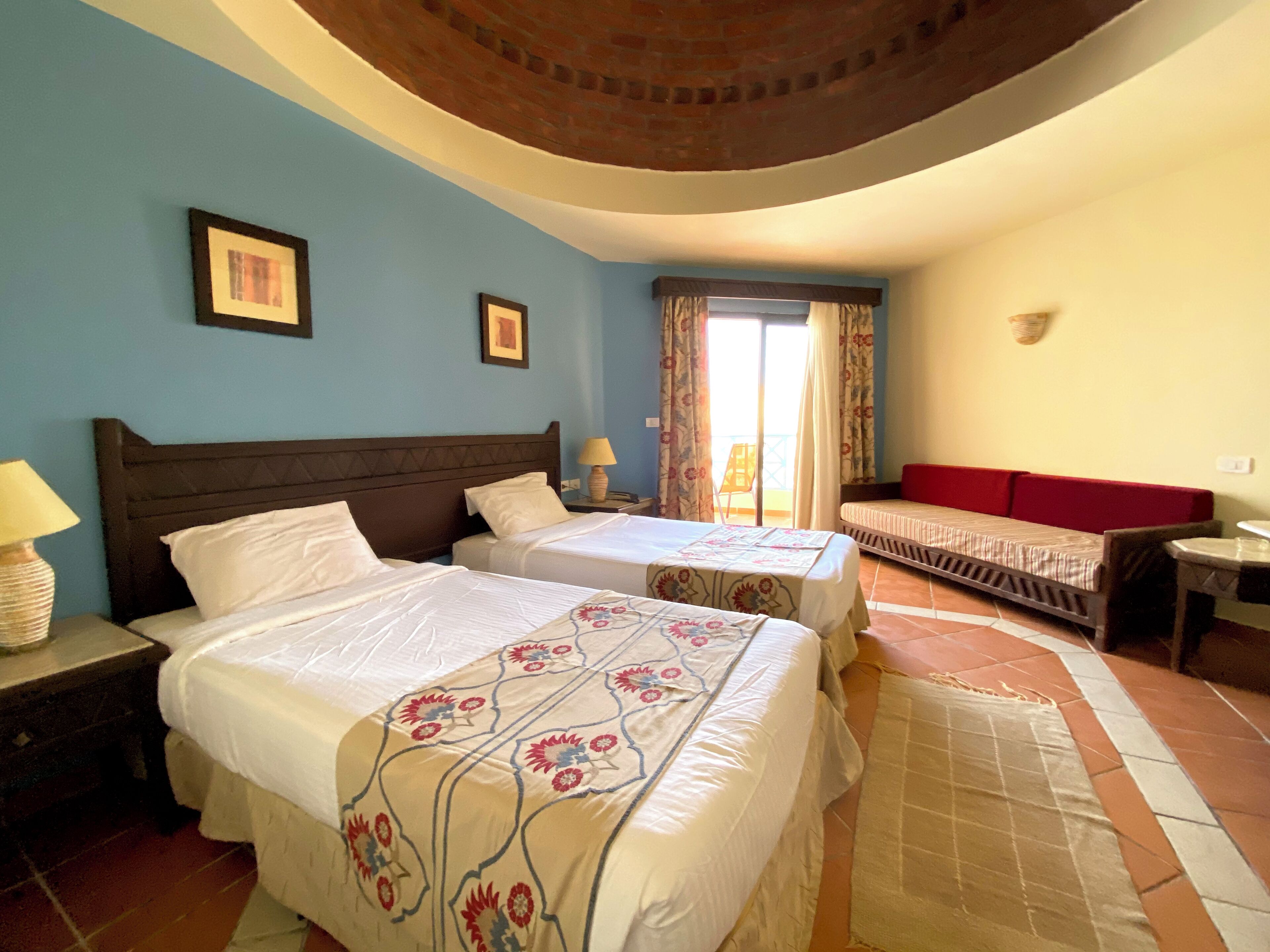 Habitación de hotel con dos camas, un sofá, una mesita y un balcón con cortinas.