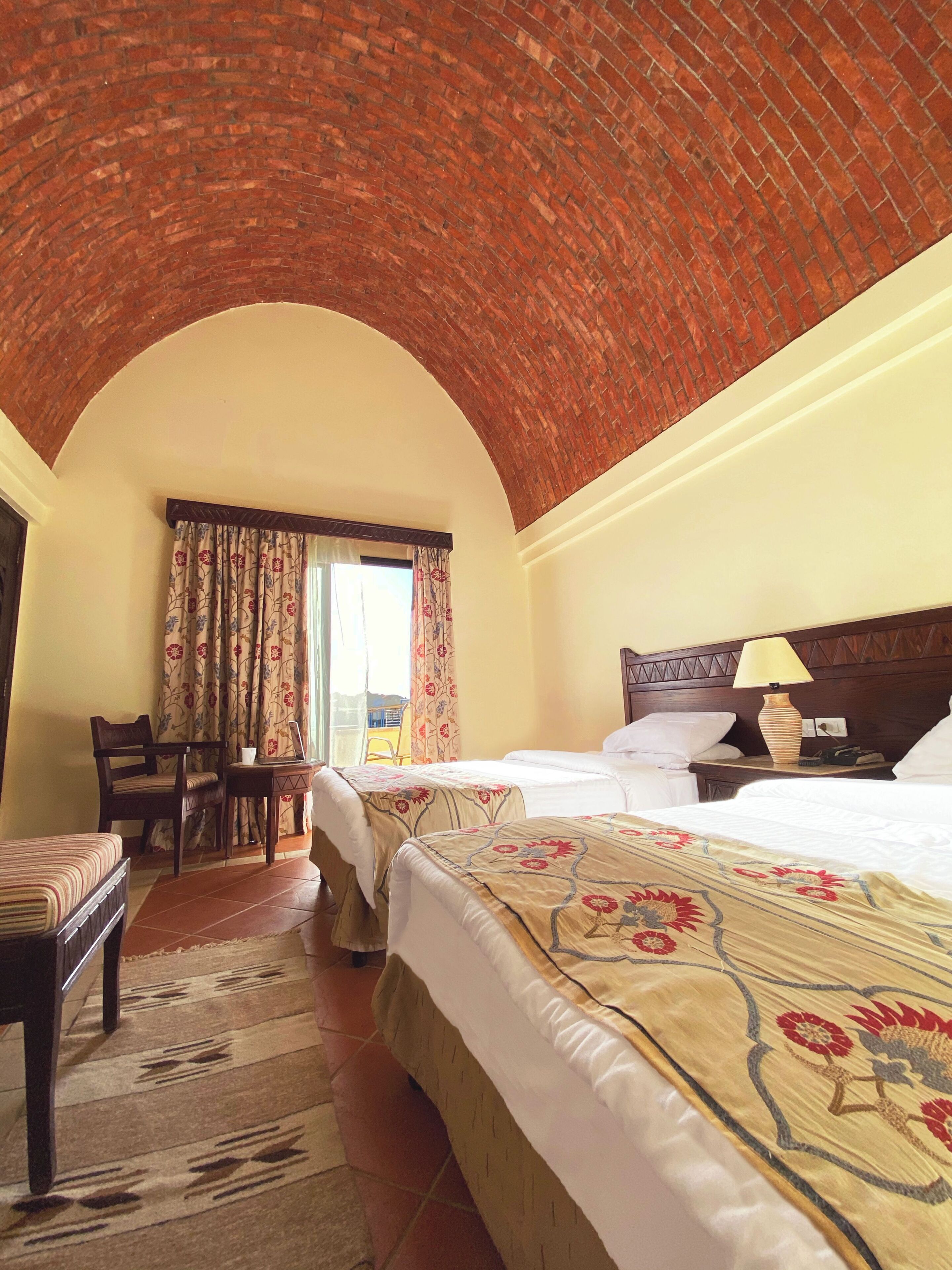 Habitación de hotel con techo de ladrillo, dos camas con colchas florales, una mesita de noche de madera, una silla y vistas al exterior.