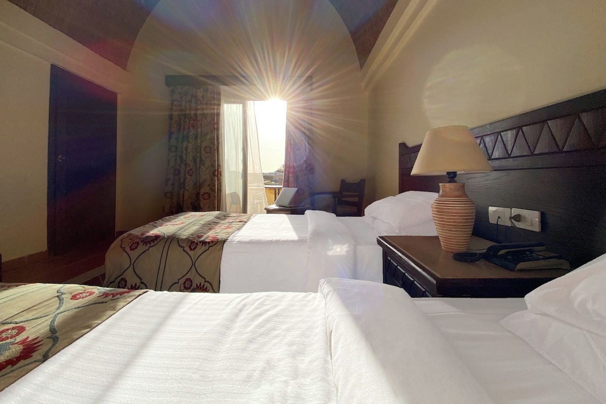 Ein Hotelzimmer mit zwei Betten, einem Schreibtisch, einer Lampe und einem fenster mit Vorhängen.