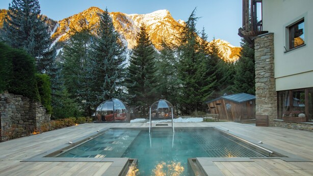 A swimming pool with a stone wall and wooden deck, surrounded by trees and a mountain in the background. - QC Hotel Monte Bianco (Pre-Saint-Didier)