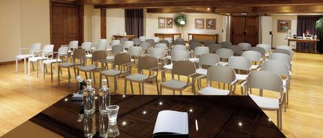 A conference room with rows of chairs, a wooden table with a notebook, glasses, and bottles, and a wooden ceiling.
