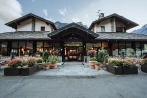 Entrada de un hotel con porche cubierto, plantas en macetas y montañas al fondo.