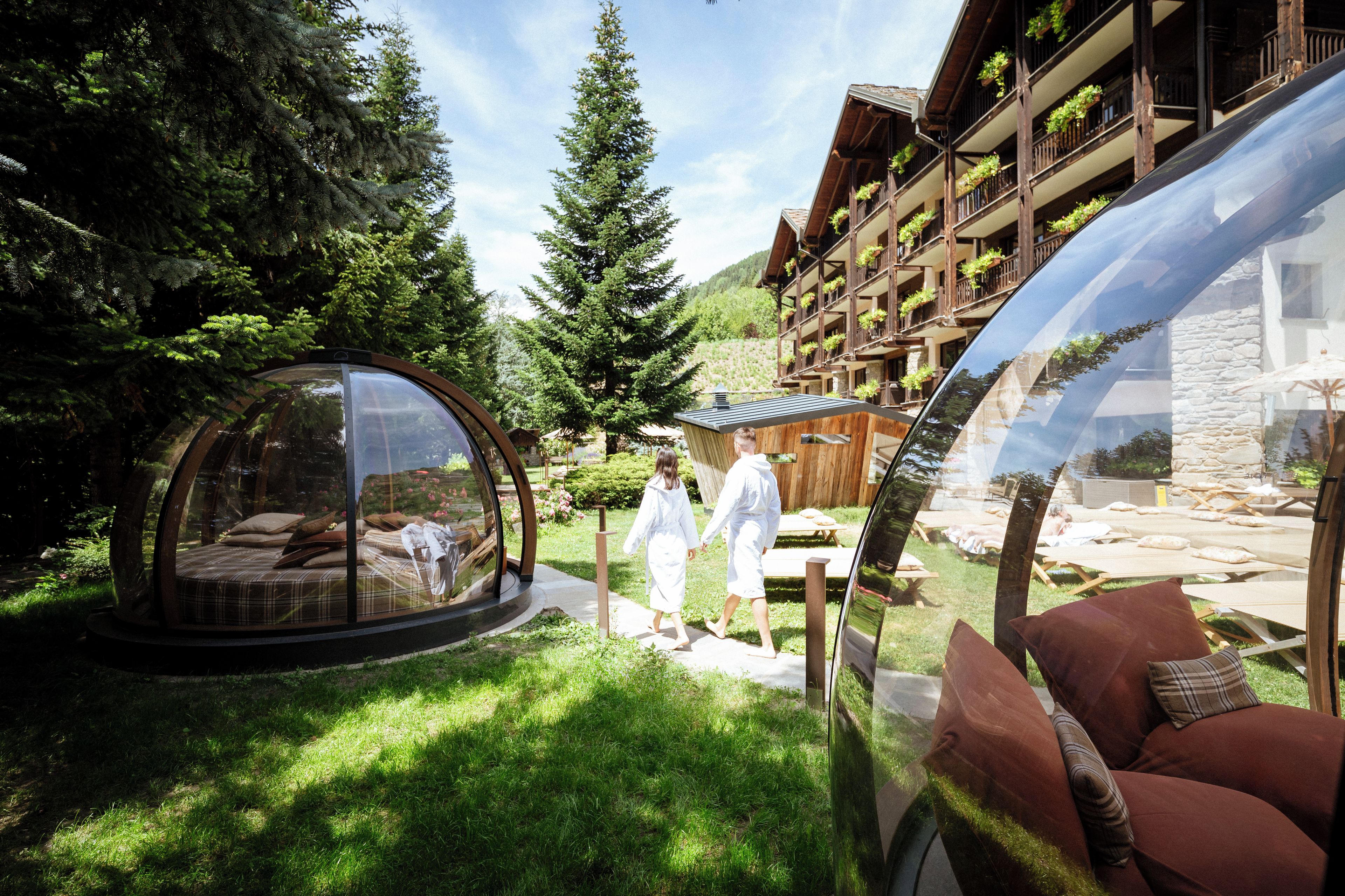 A transparent dome structure with a bed inside, surrounded by a lush garden and a wooden building with balconies.