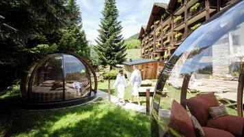 A transparent dome structure with a bed inside, surrounded by a lush garden and a wooden building with balconies.