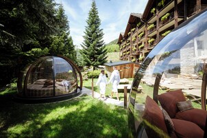 A transparent dome structure with a bed inside, surrounded by a lush garden and a wooden building with balconies.