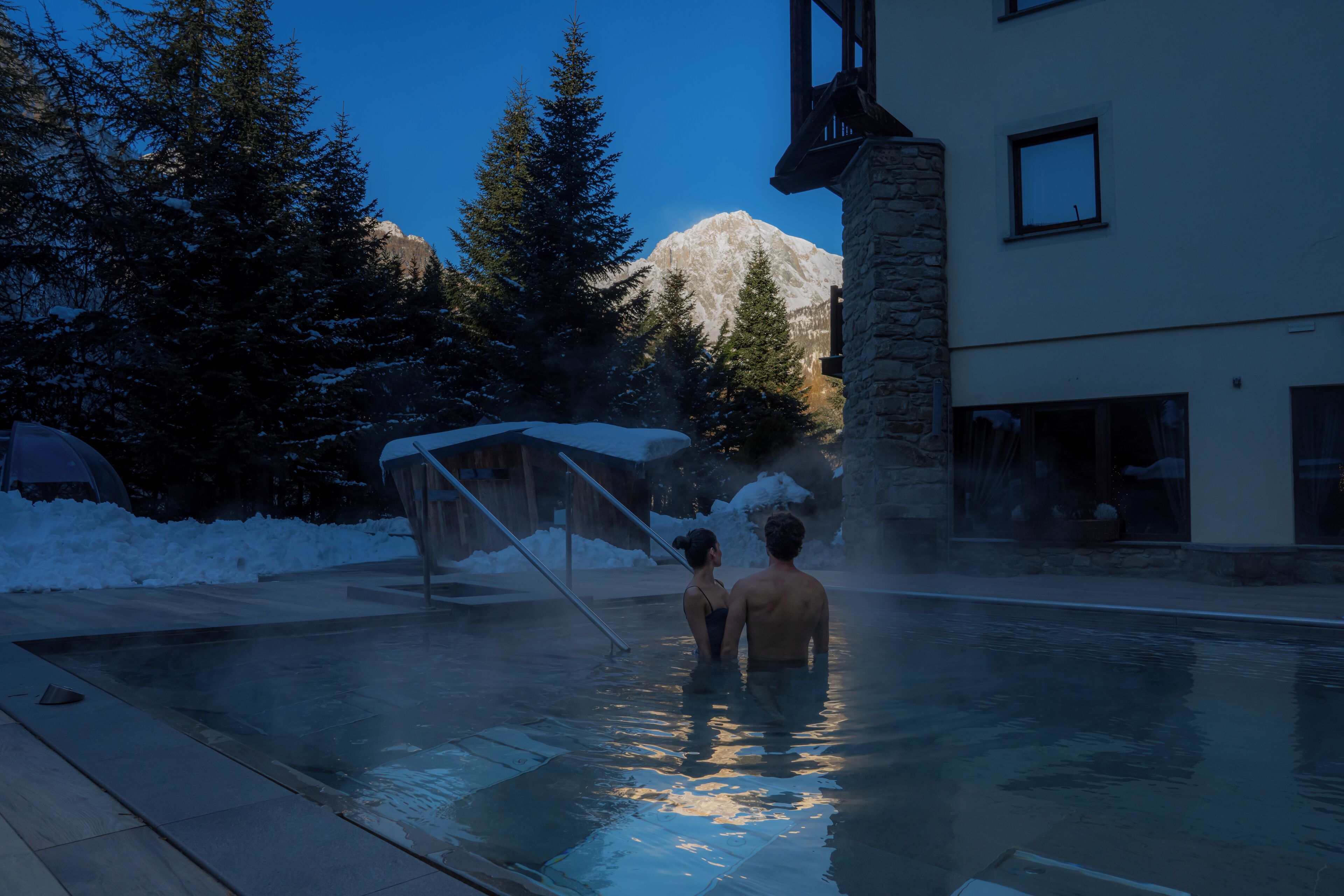 Un espace piscine avec un bâtiment en pierre, un sol recouvert de neige et une montagne en arrière-plan.
