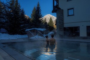 Zona de piscina con un edificio de piedra, suelo cubierto de nieve y una montaña al fondo.