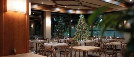 Un restaurante con mesas y sillas de madera, un árbol de Navidad y vistas de la ciudad por la noche.