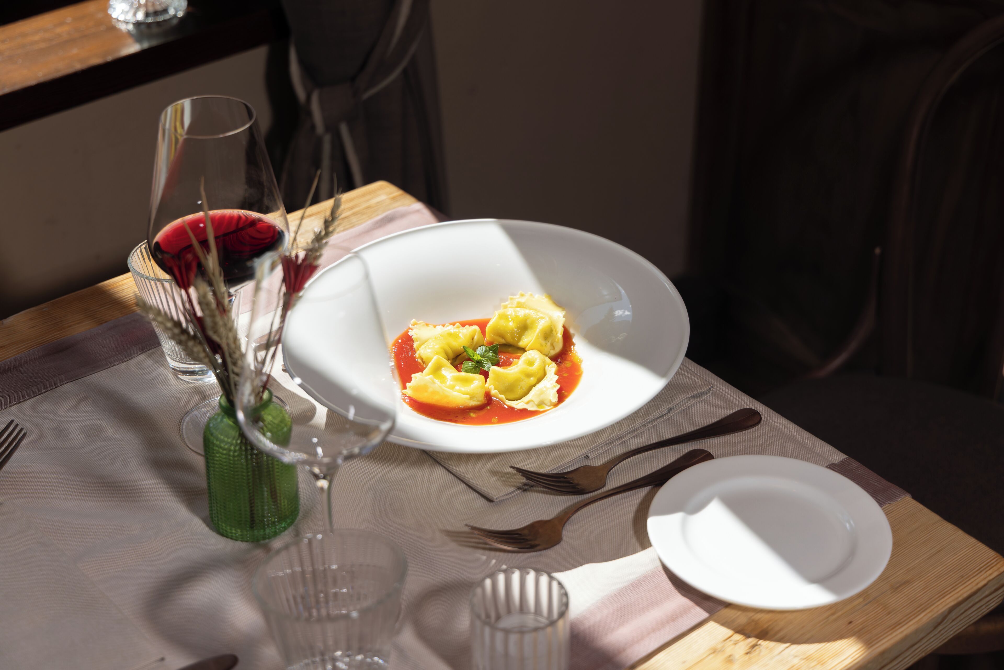 Una mesa de comedor con un plato de ravioles, un vaso de agua y un jarrón verde con flores secas.