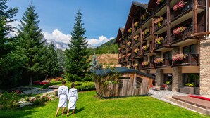 A hotel with multiple balconies adorned with flowers, a well-maintained garden, and a view of mountains in the background. - QC Hotel Monte Bianco (Pre-Saint-Didier)