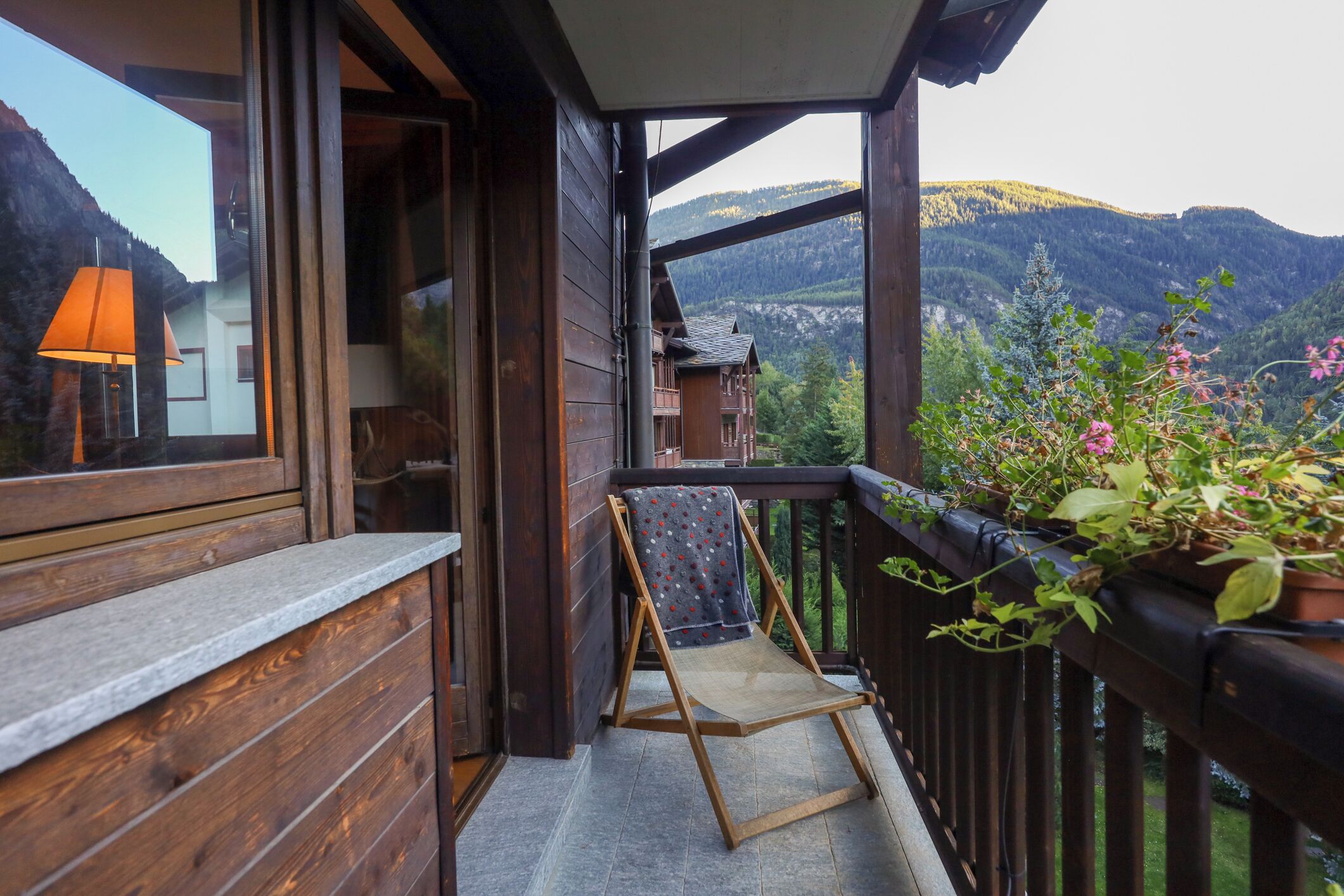 A wooden balcony with a chair, a potted plant, and a view of mountains.