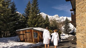 A wooden cabin with a stone wall, surrounded by snow-covered ground and trees, with a mountain in the background. - QC Hotel Monte Bianco (Pre-Saint-Didier)