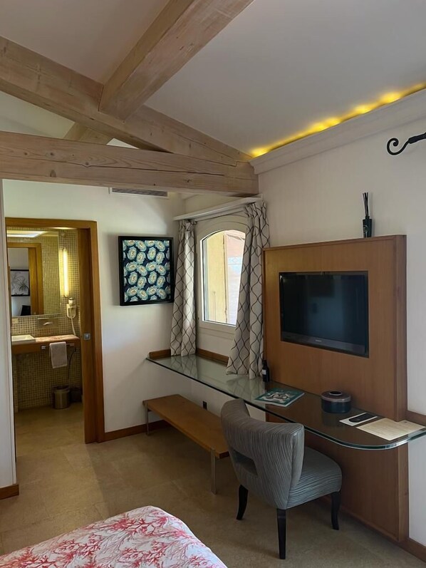 A compact bedroom with a glass desk, a television, a bed with floral bedding, a bench, and a bathroom visible through an open door. - Bastide de Ramatuelle (Ramatuelle)