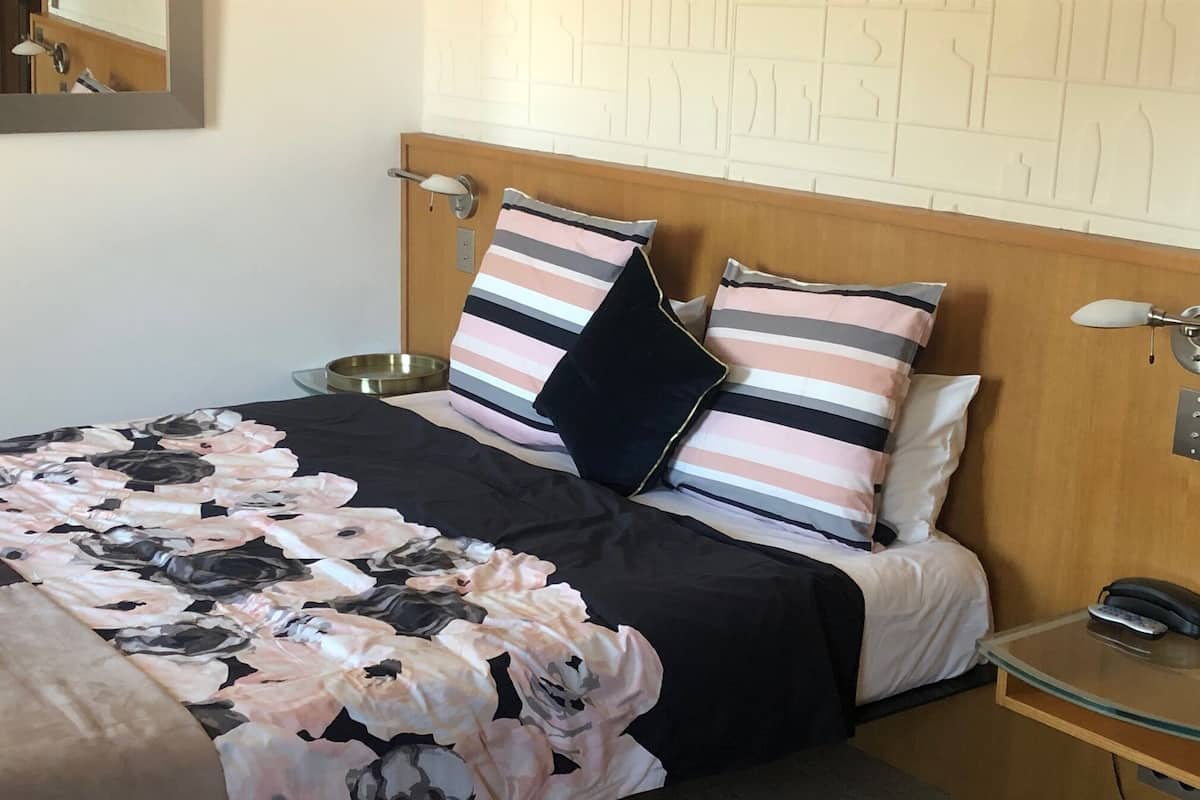 A neatly made bed with floral bedding, two striped pillows, a bedside table with a lamp, and a wall-mounted mirror.