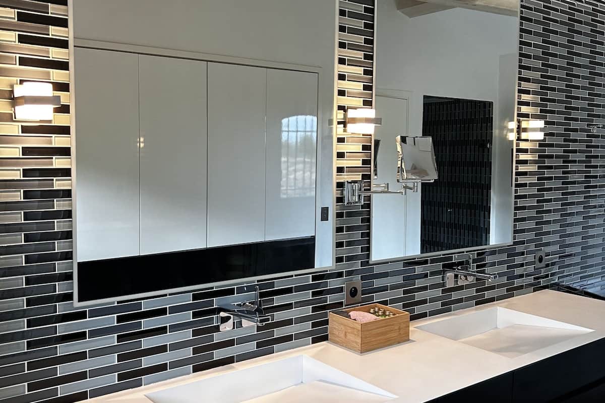 A modern bathroom with a black and white tiled backsplash, a large mirror, and a long white countertop with a wooden box.
