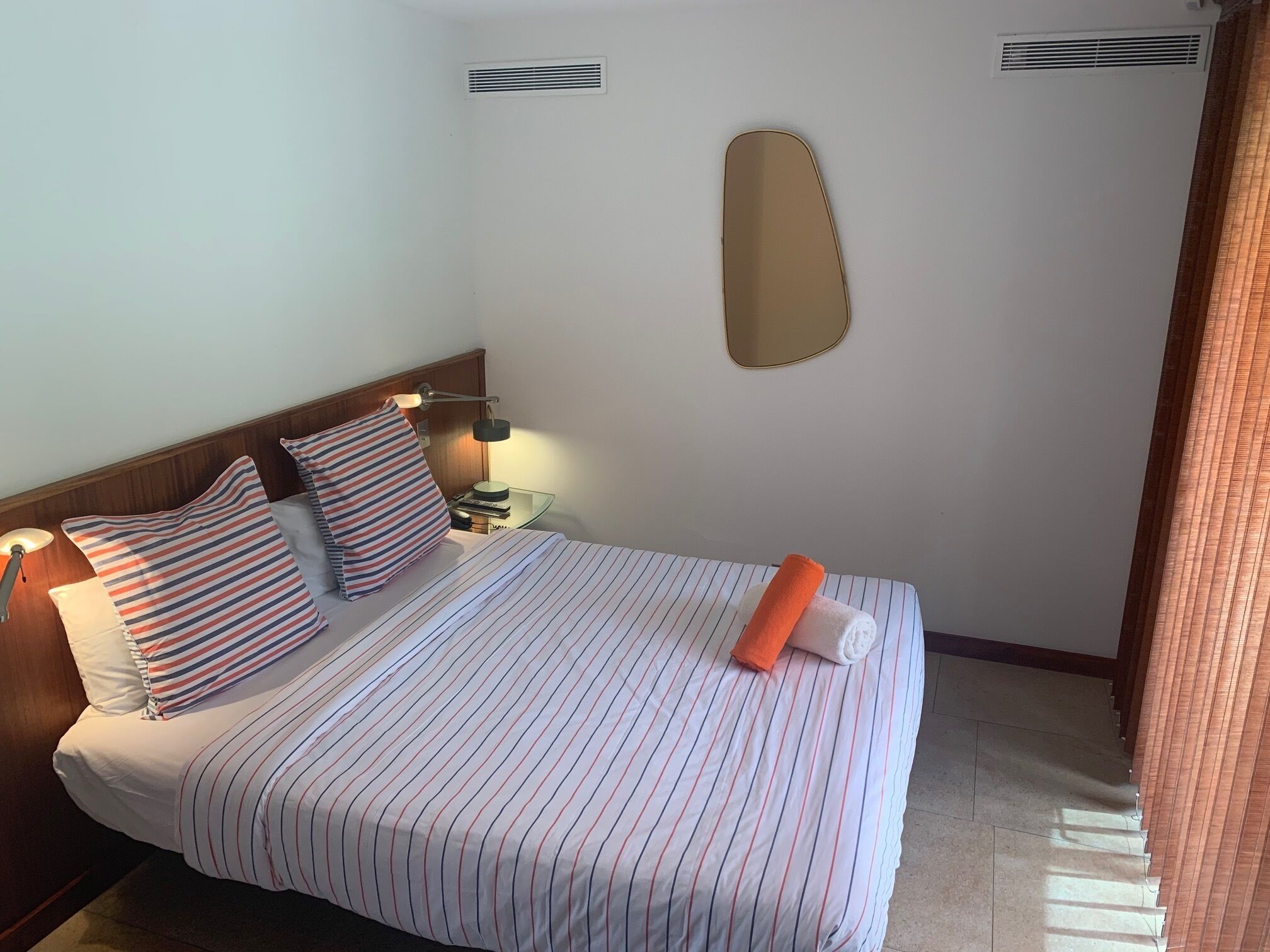 A hotel room with a bed, striped pillows, a bedside lamp, a mirror, and a window with blinds.