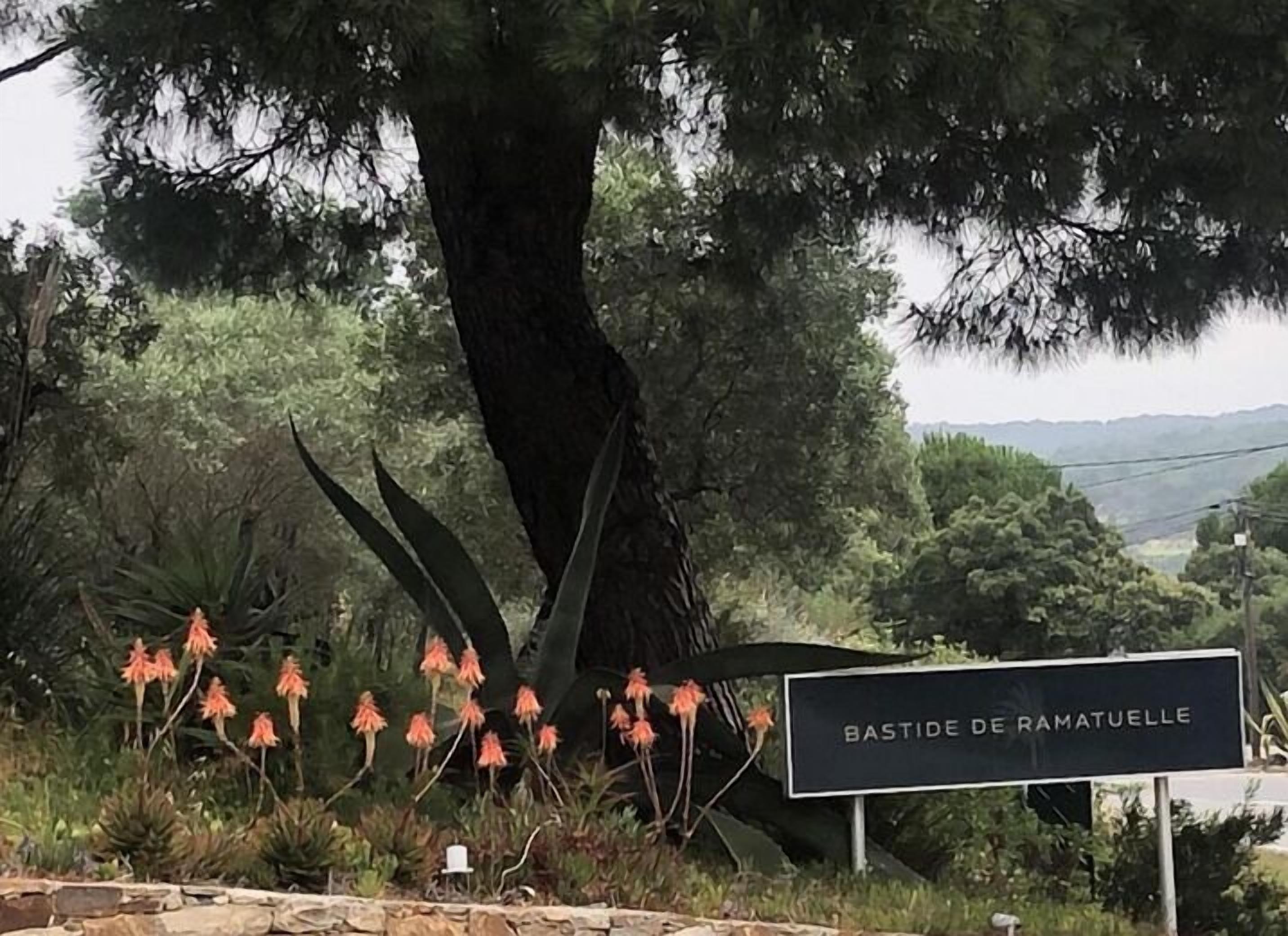 Un letrero que dice "BASTIDE DE RAMATUELLE" con un fondo de árboles y plantas florecientes.