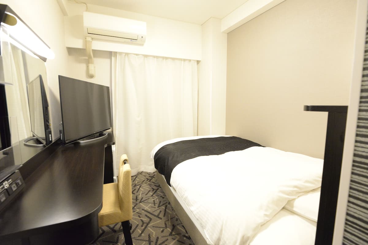 A hotel room with a bed, a desk with a computer, a chair, a TV, and an air conditioning unit.