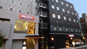 Un edificio con un letrero que indica APA HOTEL. Tiene varios pisos, una escalera y un estacionamiento con coches.