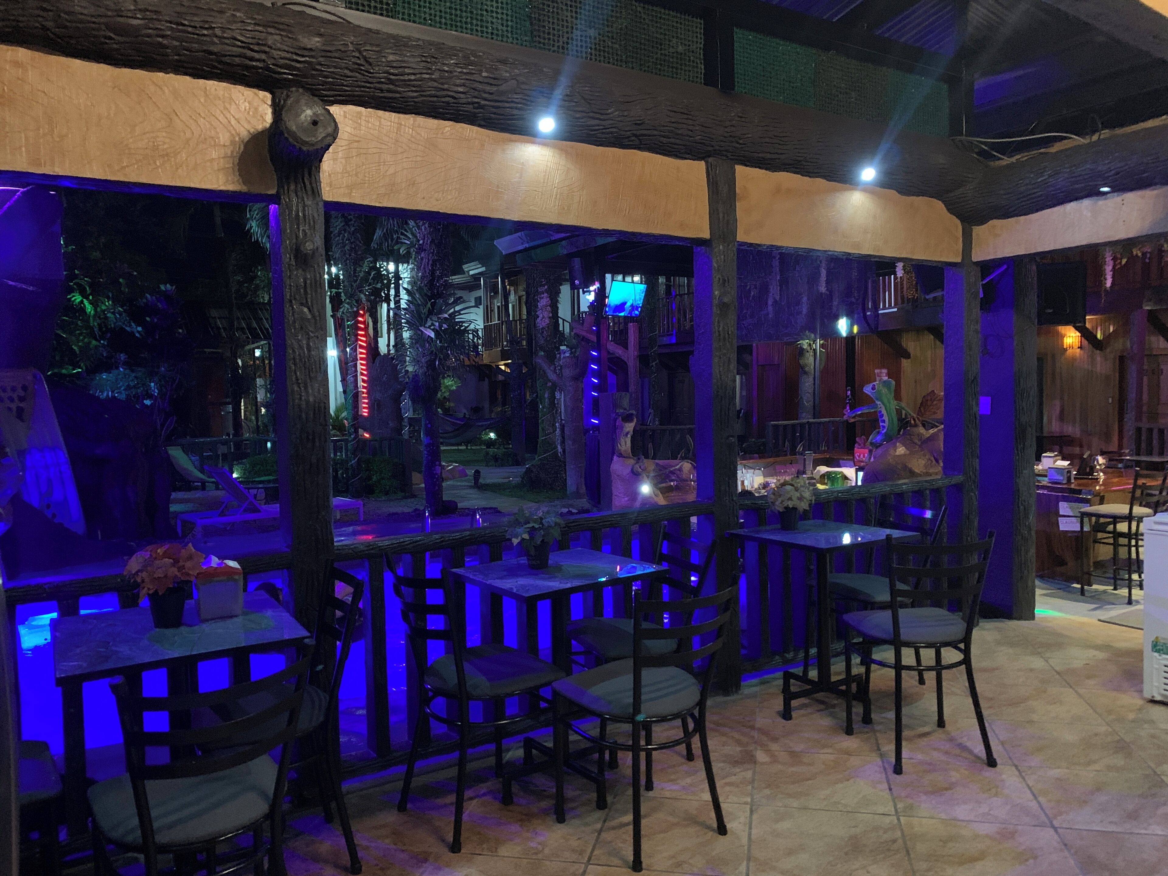 An outdoor restaurant area with wooden tables and chairs, surrounded by a wooden fence and a view of a pool area.