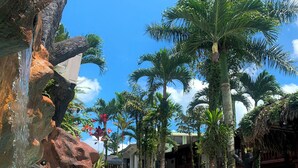 A pool with clear blue water, surrounded by palm trees and a rock formation. - Poshtel Arenal (La Fortuna)