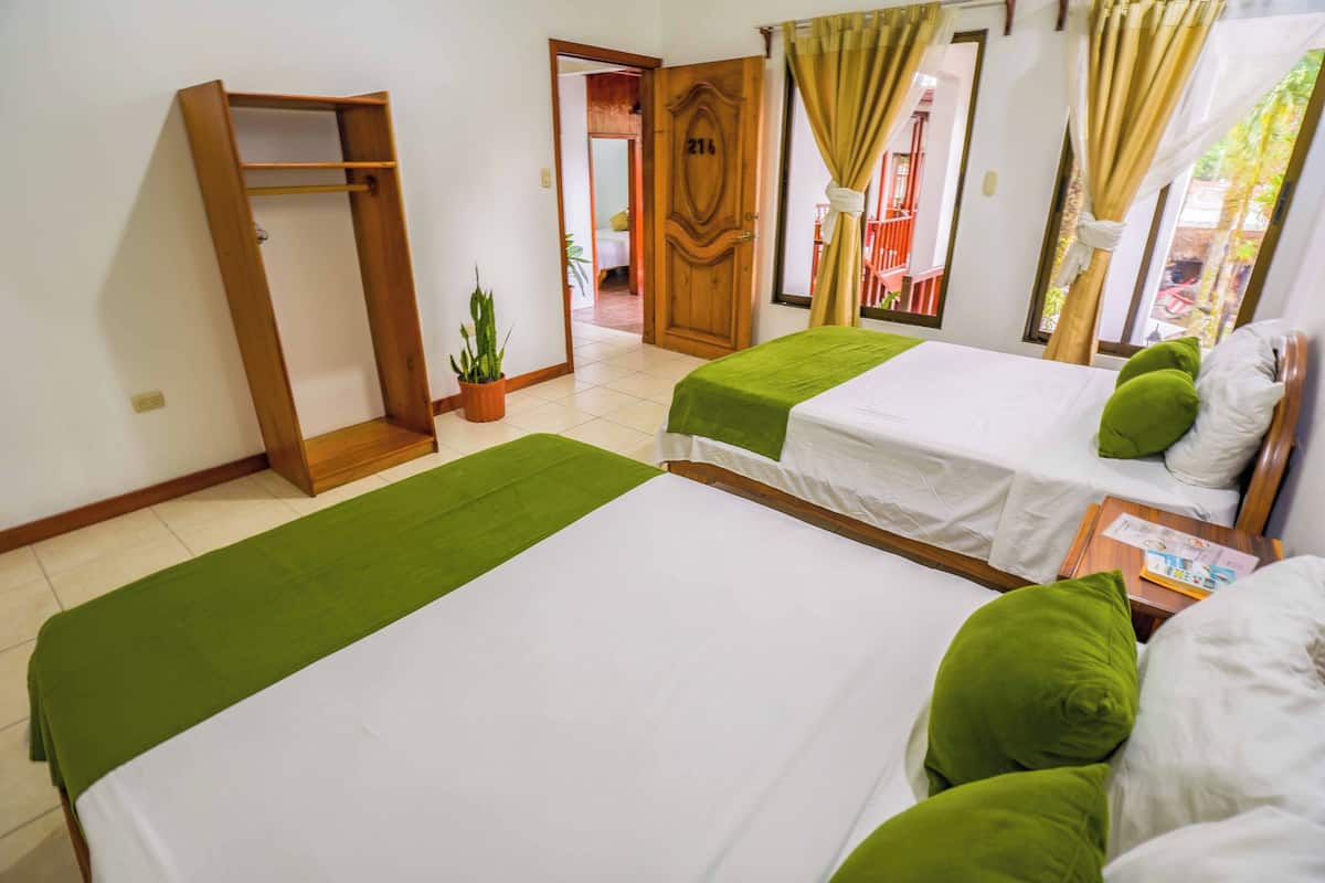 A hotel room with two beds, a wooden wardrobe, a potted plant, and a view of the outside.