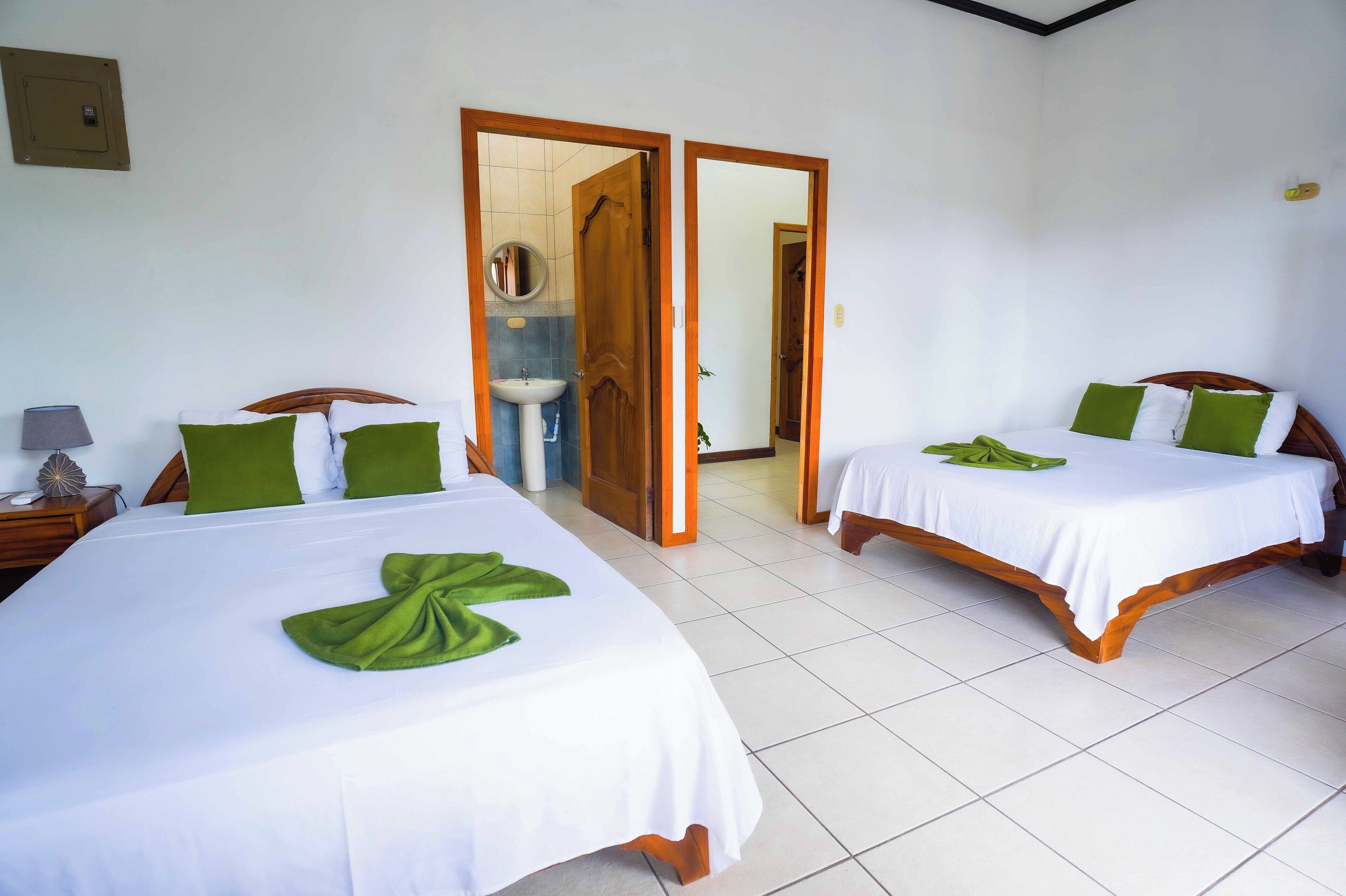 Hotelkamer met twee bedden, elk met een witte spreidek en groene kussens. Er is een houten hoofdbord, een nachtkastje met een lamp, en een badkamer is door een geopende deur zichtbaar.