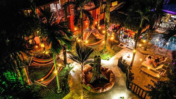 A well-lit outdoor area with a pool, hammocks, and seating areas surrounded by palm trees. - Poshtel Arenal (La Fortuna)