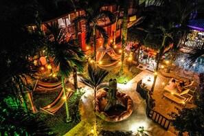 A well-lit outdoor area with a pool, hammocks, and seating areas surrounded by palm trees. - Poshtel Arenal (La Fortuna)