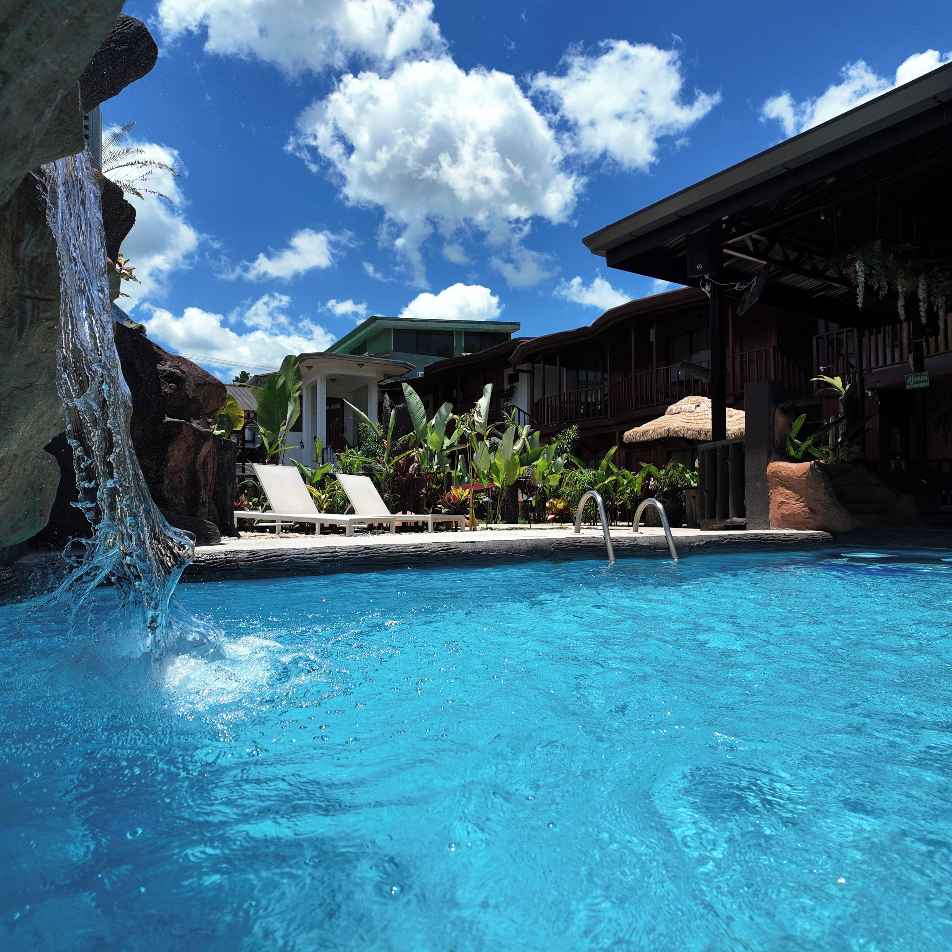 A swimming pool with clear blue water, surrounded by lounge chairs, a waterfall feature, and a building with a green roof.