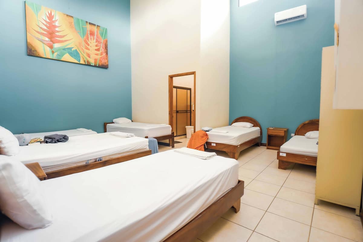 A hotel room with four single beds, a wooden headboard, a wall-mounted air conditioner, and a painting above the beds.