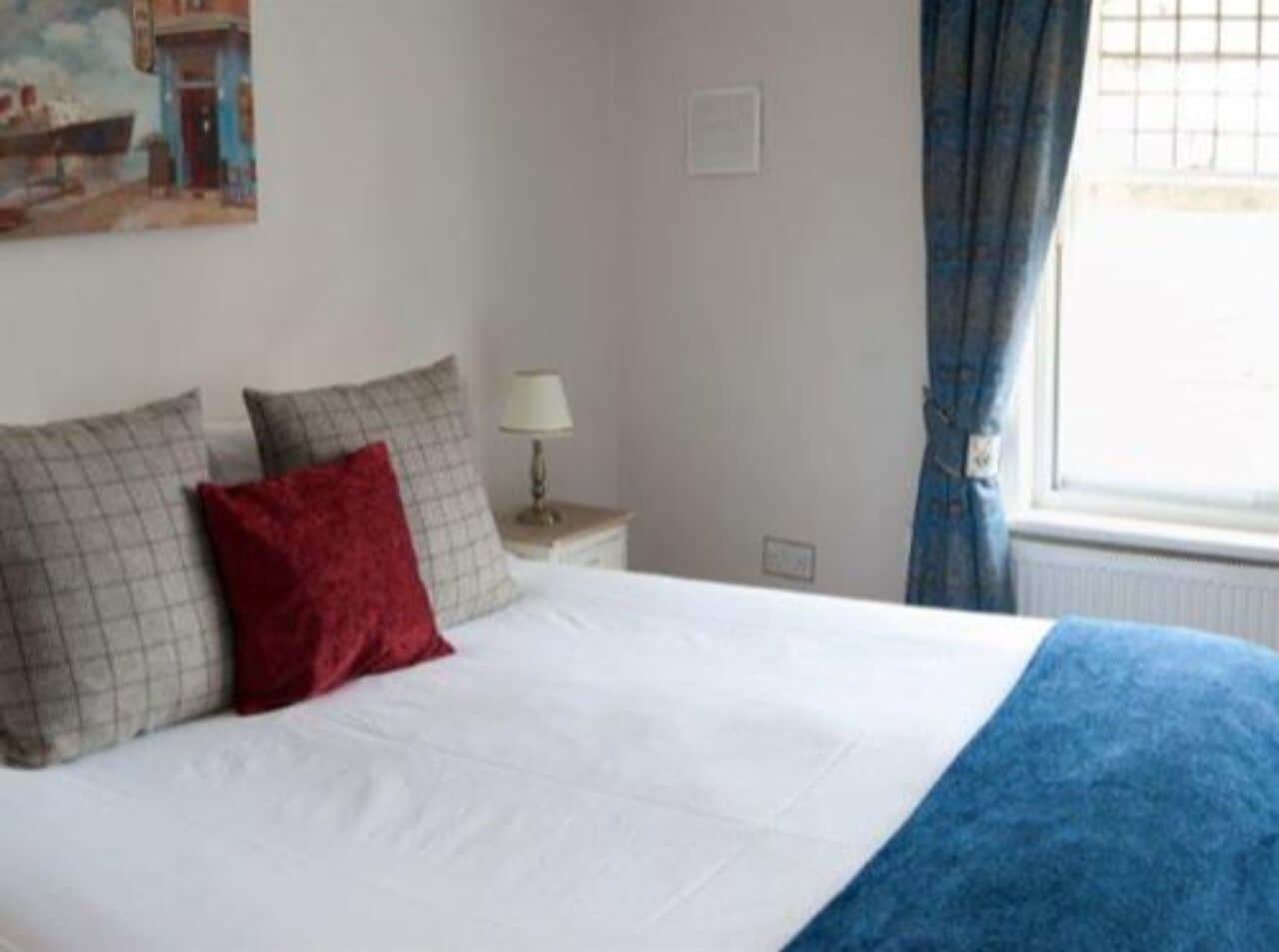 A neatly made bed with white bedding and a blue throw blanket, a small bedside table with a lamp, and a painting on the wall.