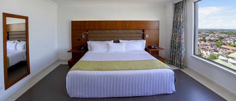 Habitación de hotel con una cama grande, cabecera de madera, dos mesitas de noche y vistas a un paisaje urbano a través de un ventanal.
